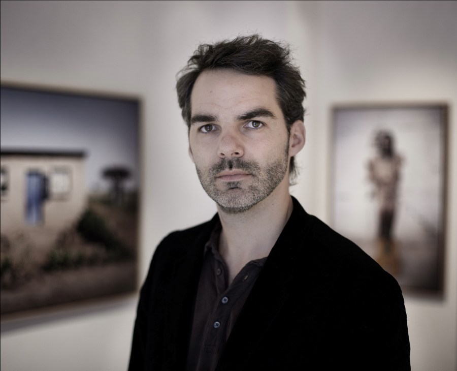 Serge Macia, founder of the Imaginaid Gallery, stands in an art gallery space with artworks visible in the background.