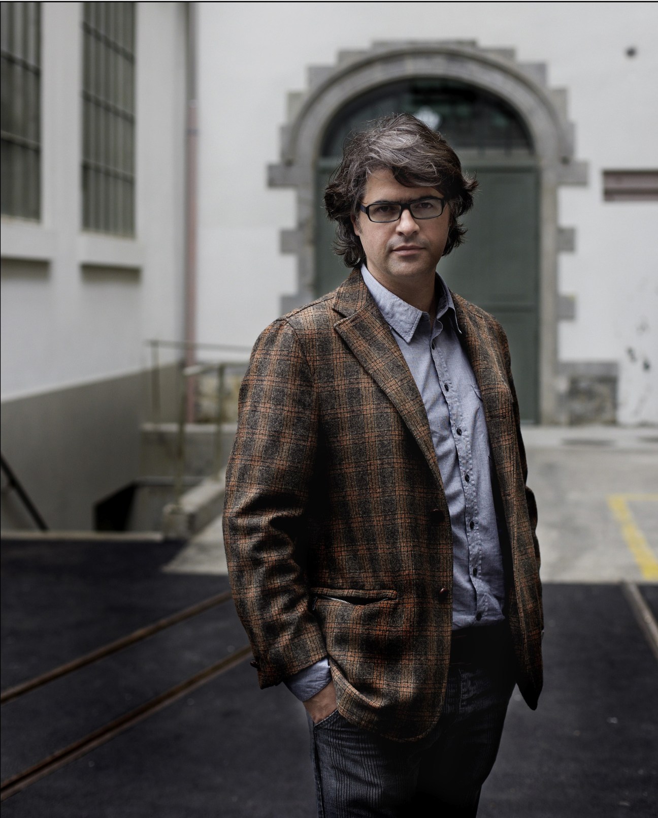 Architect Andrés Ojeda stands in a courtyard wearing a brown plaid blazer and blue shirt.