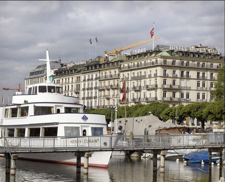 White ferry boat docked at Geneva waterfront with Hotel de la Paix and Rolex building in background.