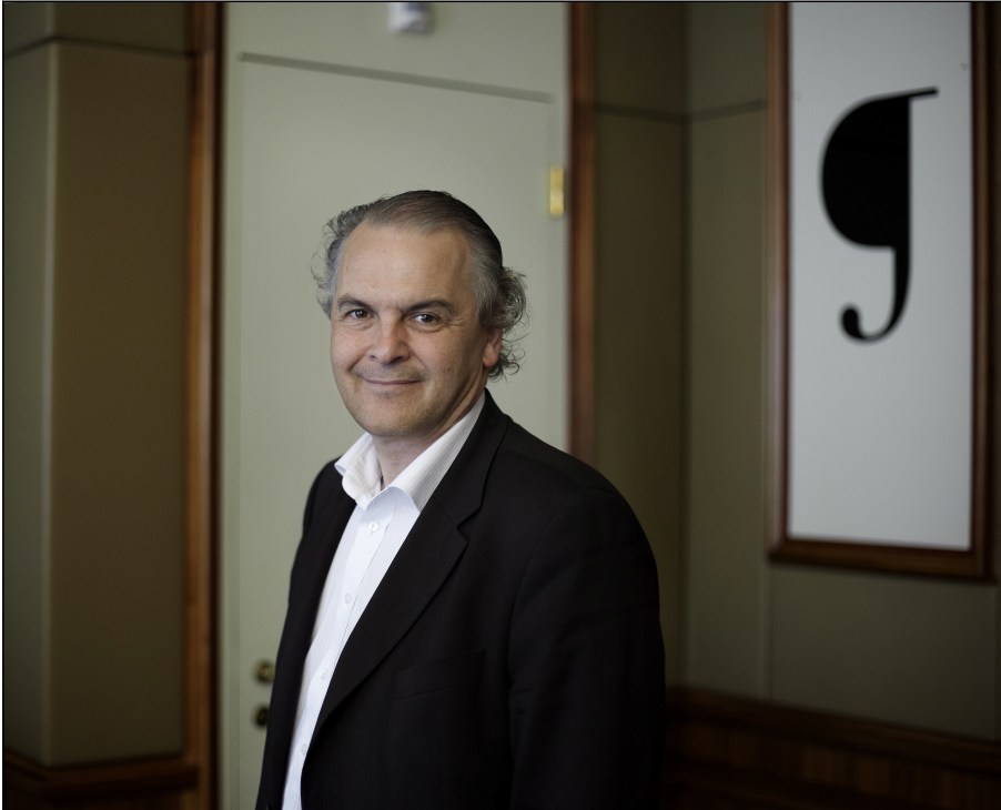 Rémy Pagani, mayor of Geneva, poses in a modern corridor wearing a black blazer and white shirt.