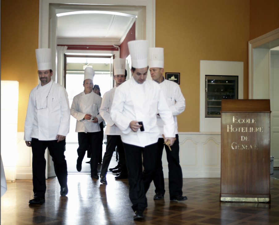 Professional chefs in white chef coats and tall white hats walking through a hallway at École Hôtelière de Genève.
