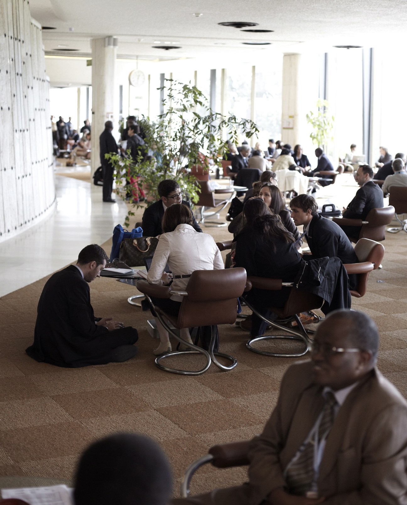 UN staff members take a break in the cafeteria, sitting in groups on brown chairs throughout the modernist space.