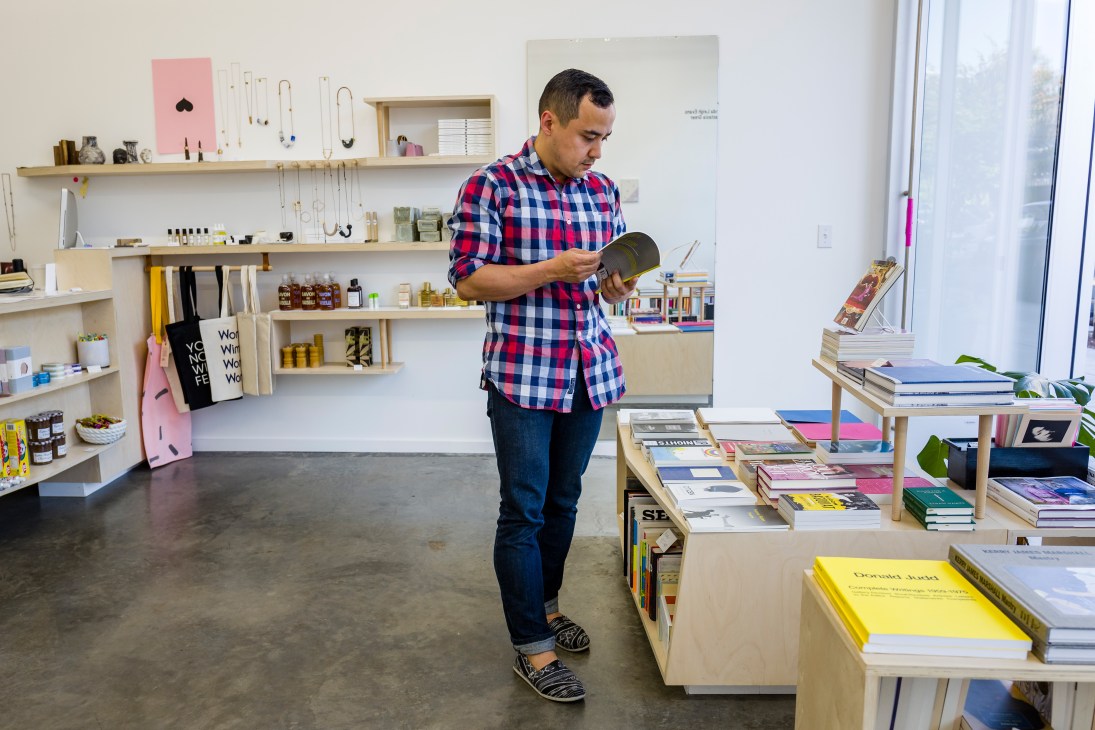 Man in red plaid shirt browsing books and products at a minimalist retail shop with wooden shelving and natural light.