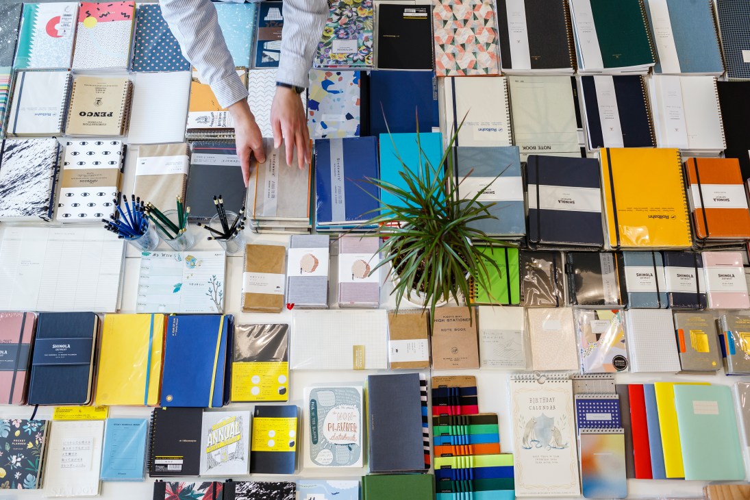 Overhead view of colorful notebooks and stationery arranged on white surface with a hand reaching toward items.