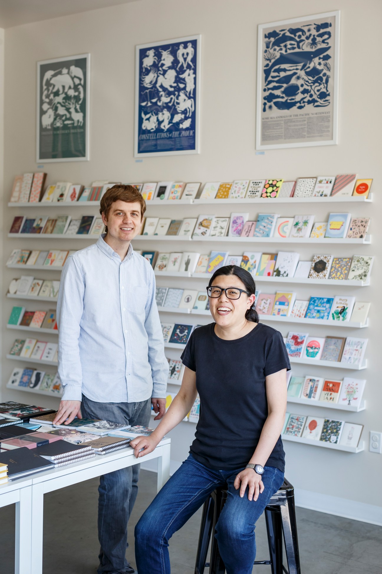 Two people pose in front of shelves displaying colorful notebooks and stationery at Little Otsu in Portland.
