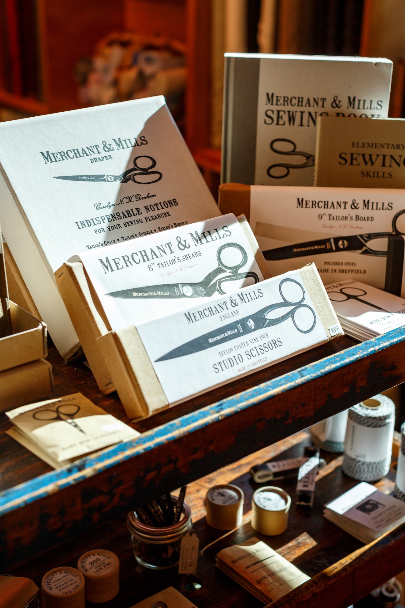 Merchant & Mills sewing products including scissors, thread, and instructional books displayed on wooden shelf.