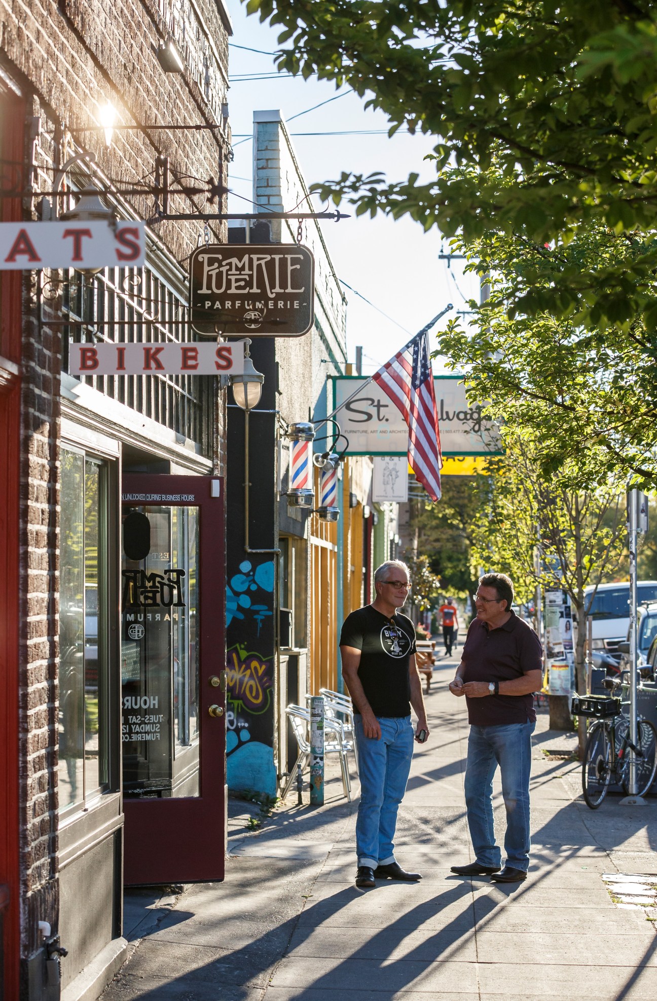 Two men standing on Division Street in Portland with storefronts including a parfumerie and barber shop visible behind them.