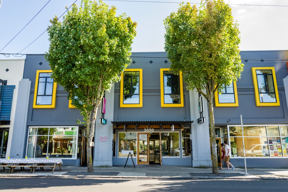 Modern storefront with yellow-framed windows, ivy-covered facade, and street-level retail shops on Division Street Portland.