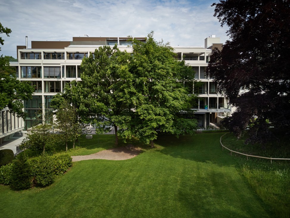Modern white apartment building with large windows overlooking a manicured park with green lawn and mature trees.