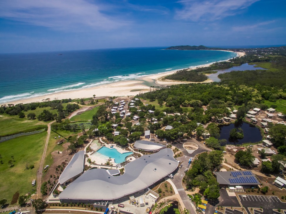 Aerial view of Elements of Byron Bay resort with curved architecture, pool, and beach nestled among rainforest.