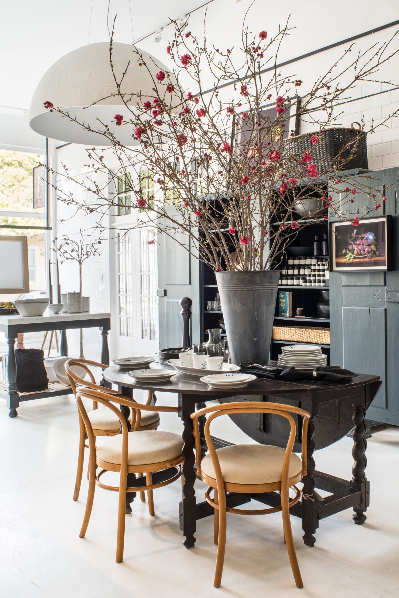 Dining room display with black table, wooden chairs, pendant lamp, and tall floral arrangement in metal vessel.