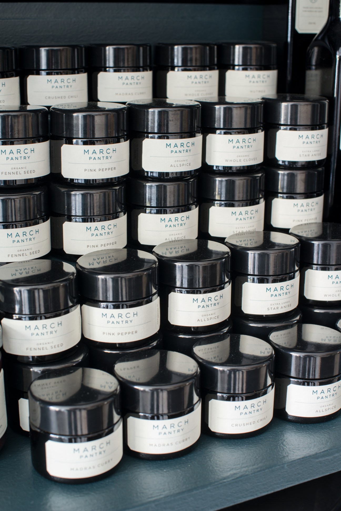 Multiple jars of March Pantry spices with black lids and cream labels arranged on a dark surface.
