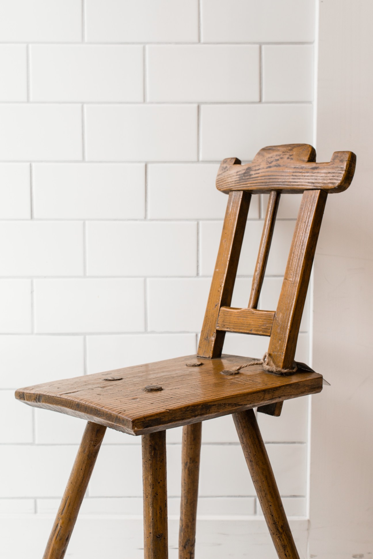Wooden chair with carved seat and backrest against white subway tile wall.