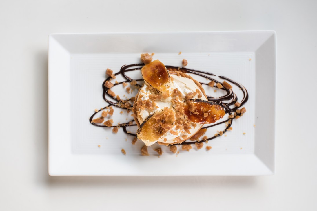Banoffee tart with caramelised banana, whipped cream, toffee crumble and chocolate drizzle on a white plate.