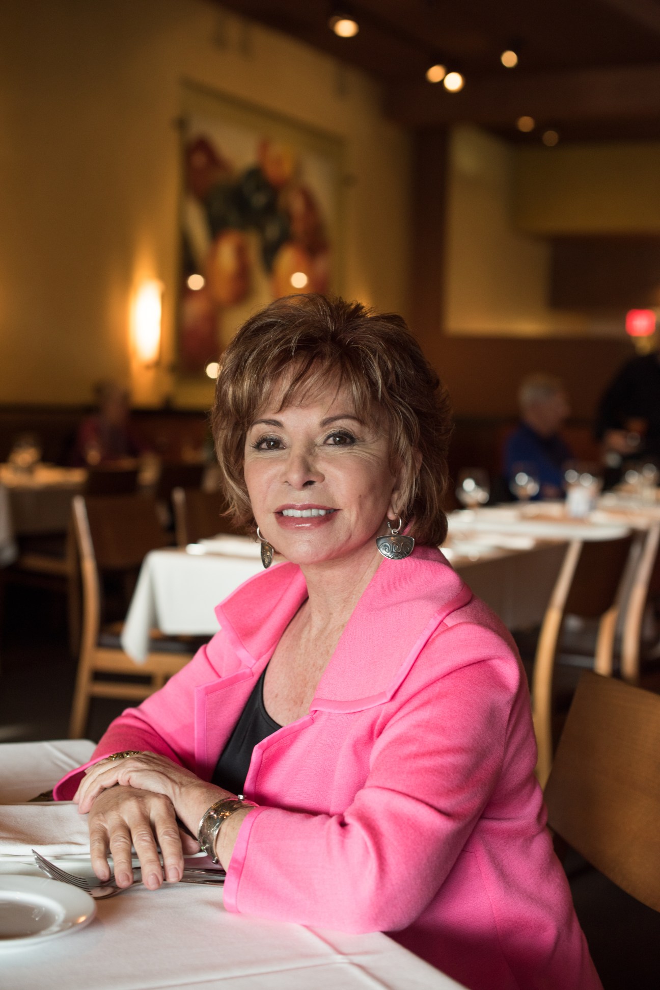 Isabel Allende seated at a restaurant table in a bright pink blazer, smiling at the camera.