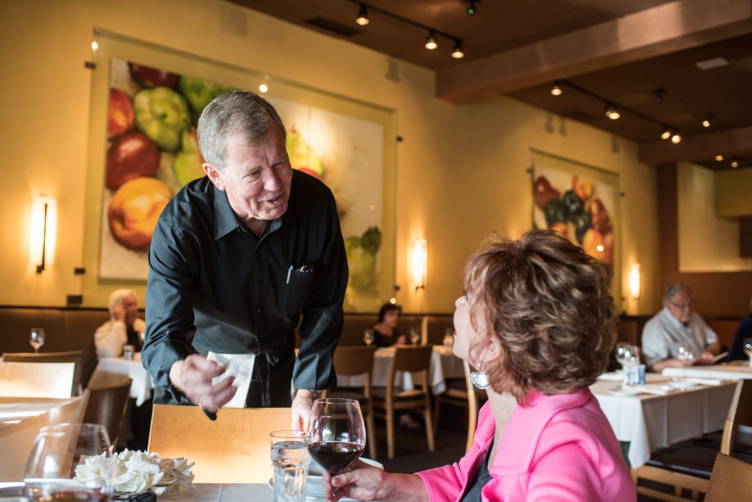 Isabel Allende in a bright pink top sits at a restaurant table with a glass of red wine, speaking with waiter Brian Weirum in a dark shirt.