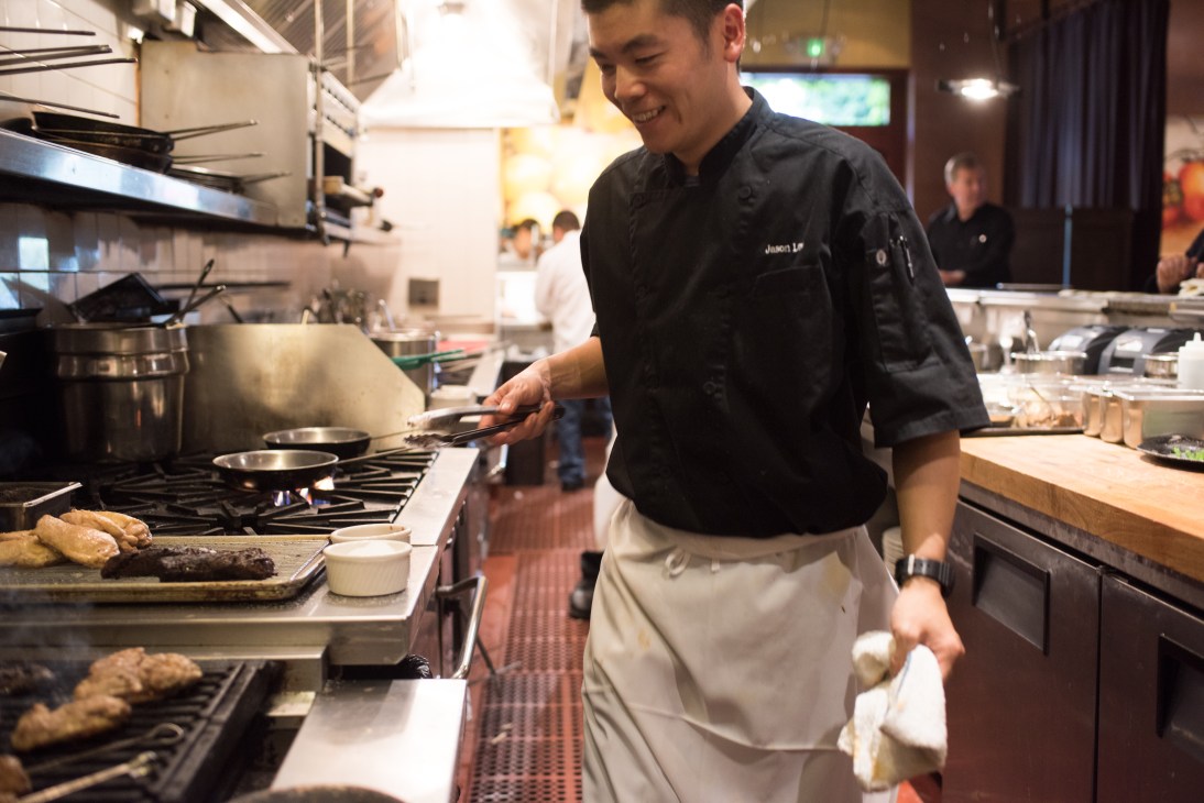 Chef working at a stove in an open kitchen with visible cooking equipment and ingredients.
