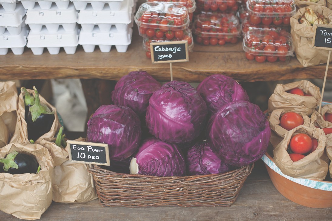 Farmers market display of fresh organic produce including purple cabbage, eggplant, tomatoes and other vegetables with handwritten price labels.
