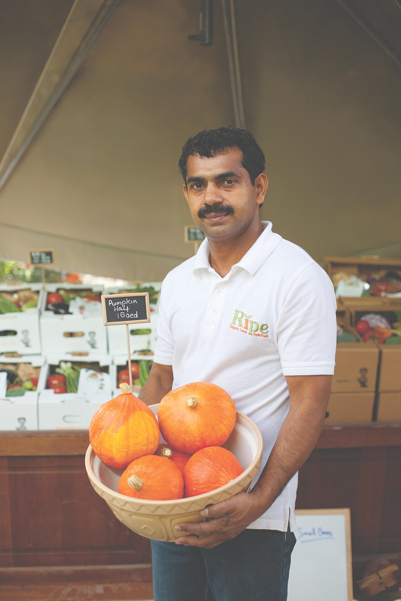 Man in white Ripe polo shirt holding basket of orange pumpkins at farmers market.