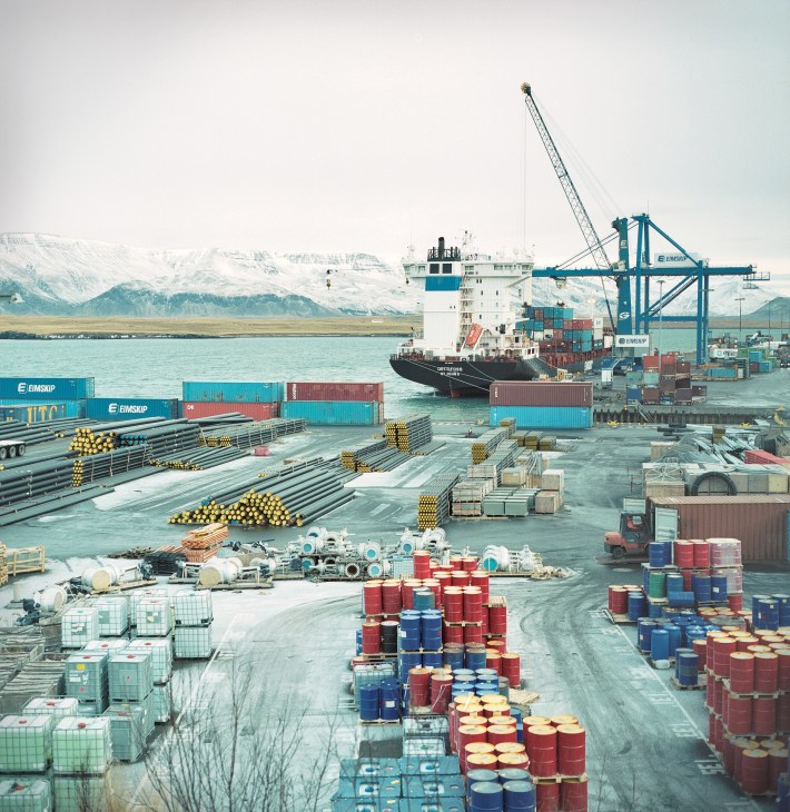Container port in Reykjavik with cargo ship, stacked containers, barrels, and snow-capped mountains in background.