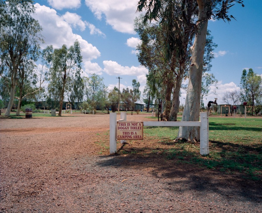 Sign at Auski roadhouse clarifying camping area is not a dog toilet, with eucalyptus trees and buildings in background.