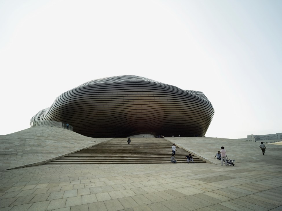 Asymmetrical dome museum with rippled metal exterior and wide plaza entrance in Ordos, China.