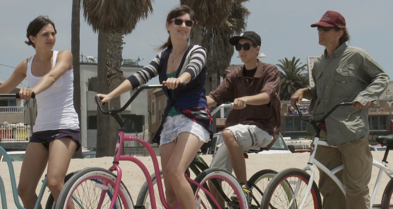 Five people on bicycles at a beach with palm trees in the background.