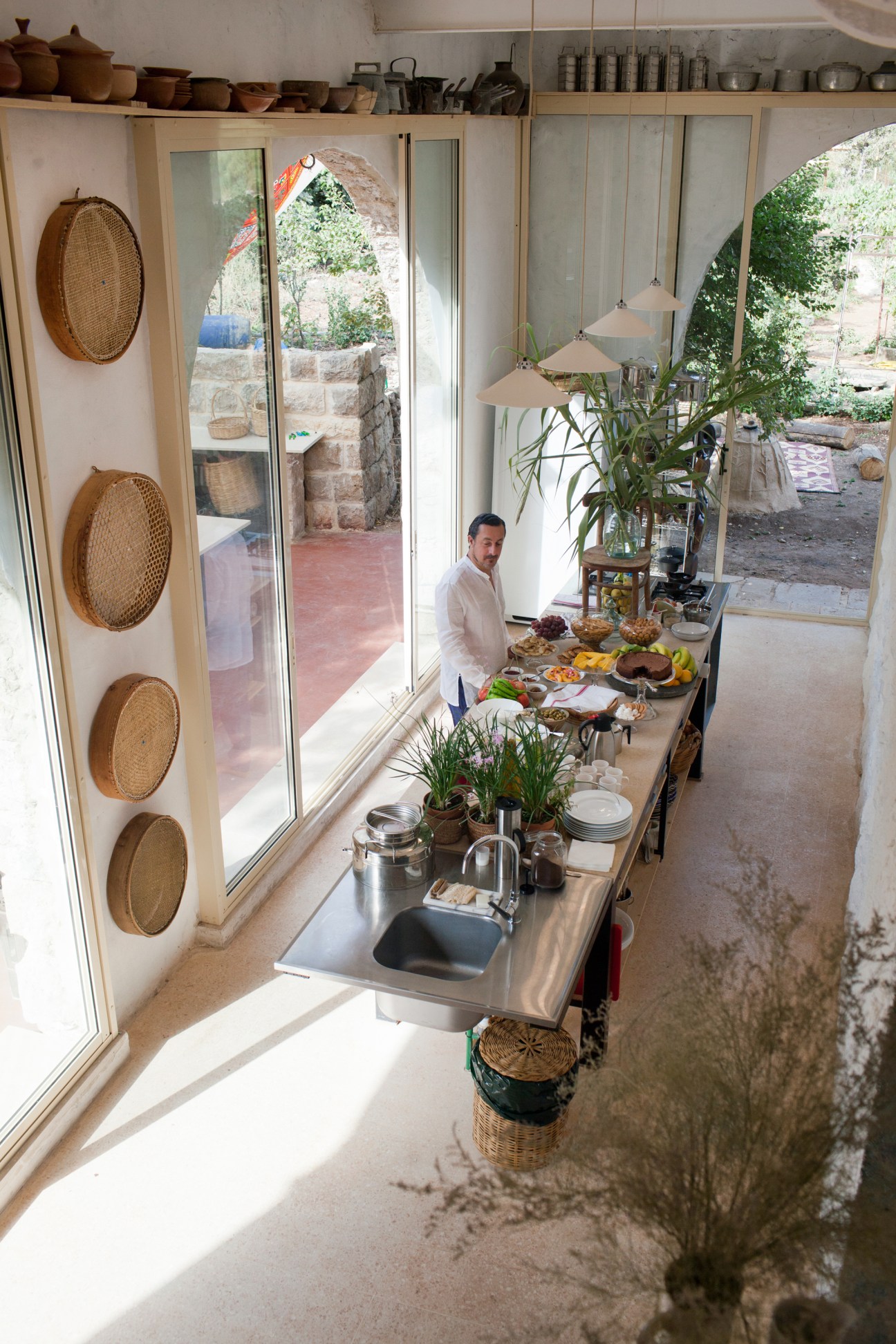 Chef Kamal Mouzawak stands at a kitchen counter with fresh fruit and food displayed, framed by large windows overlooking a stone courtyard.