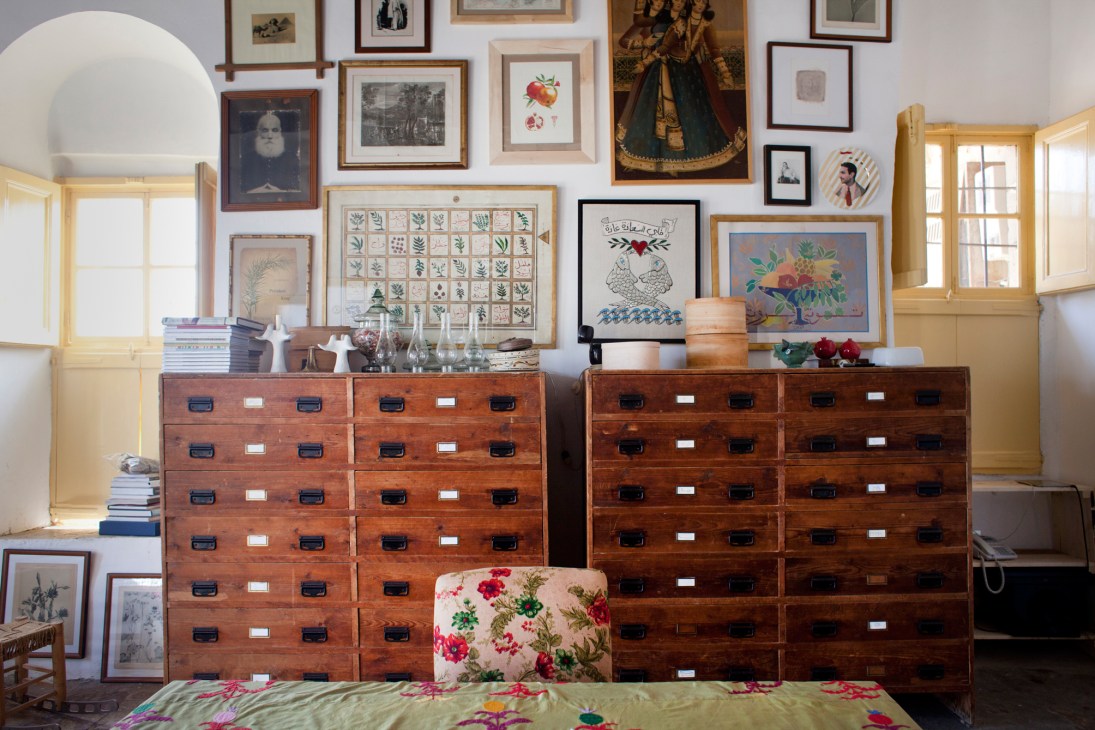 A bright library room with wooden dressers, framed artwork gallery wall, yellow-framed windows, and antique furnishings.