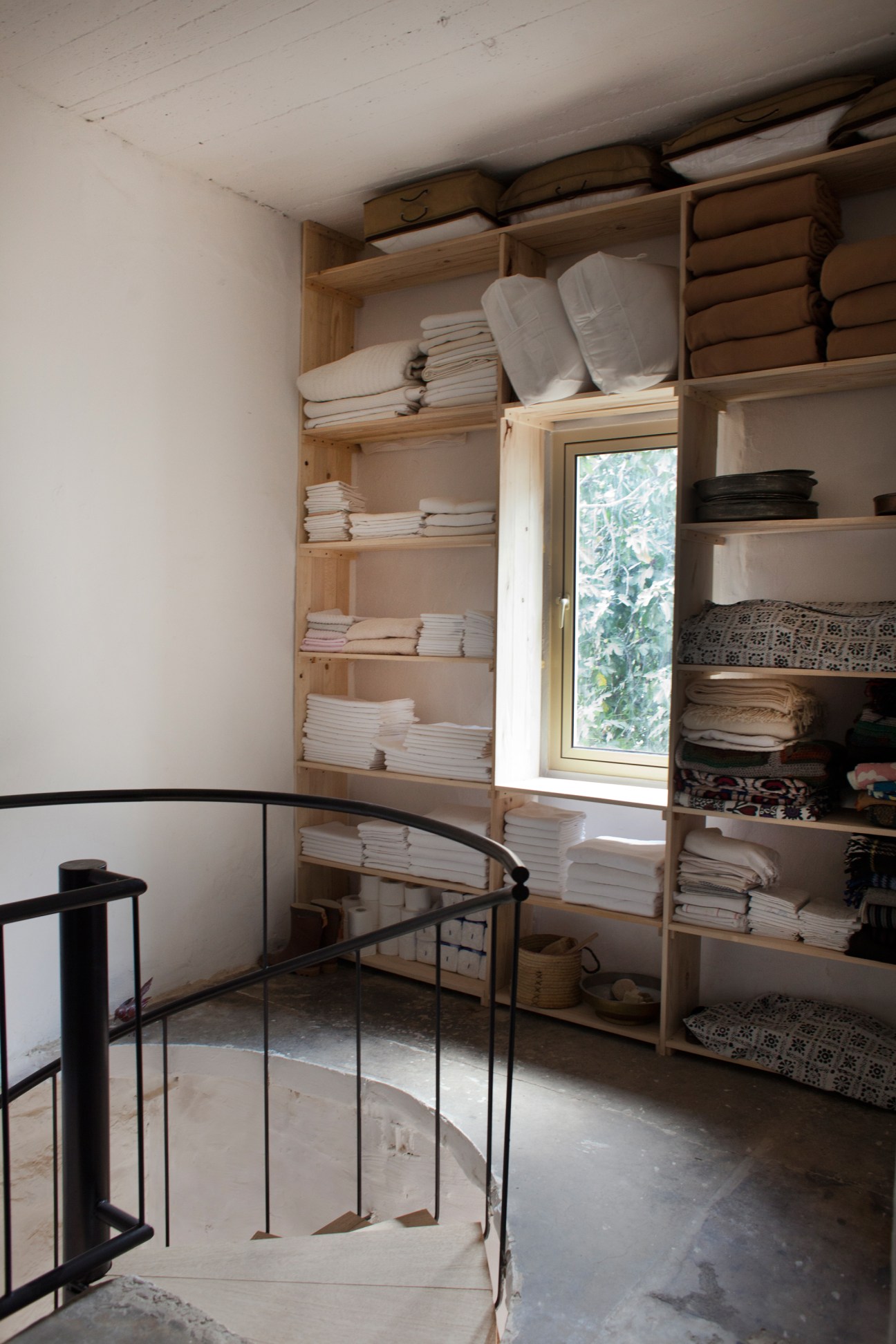 Open wooden shelving displays neatly folded white linens and textiles in a bright room with a window and modern black metal staircase.
