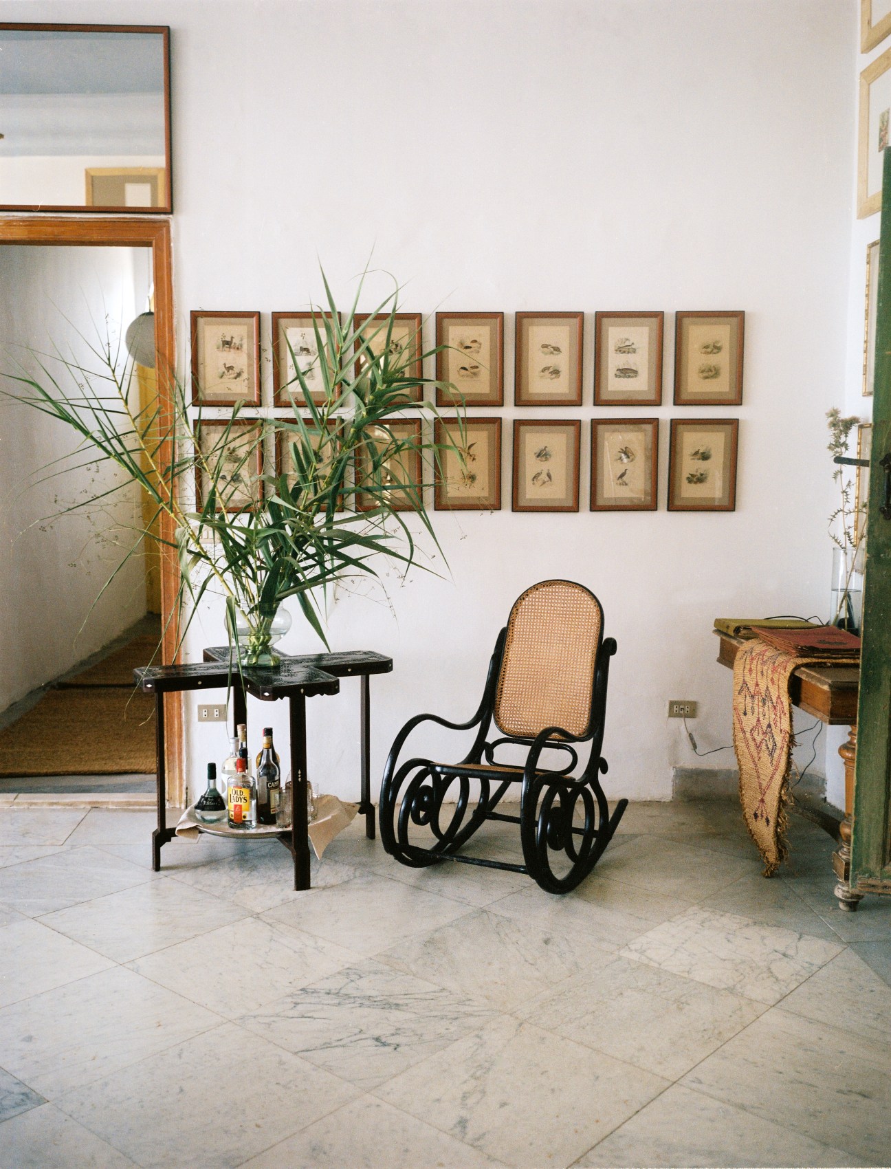 A minimalist interior with a black bentwood rocking chair, potted plant, framed botanical prints, and wooden mirror in Beit Douma.