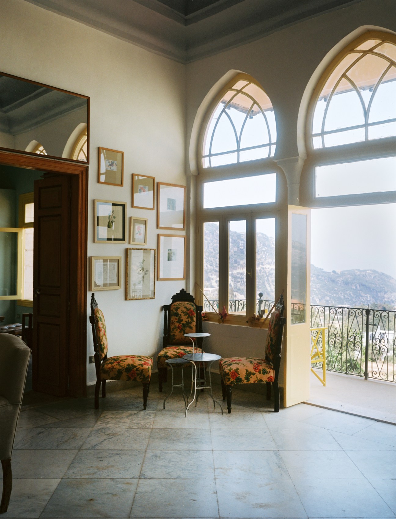 Sunlit interior room with arched windows overlooking mountains, featuring patterned chairs and framed artwork on walls.