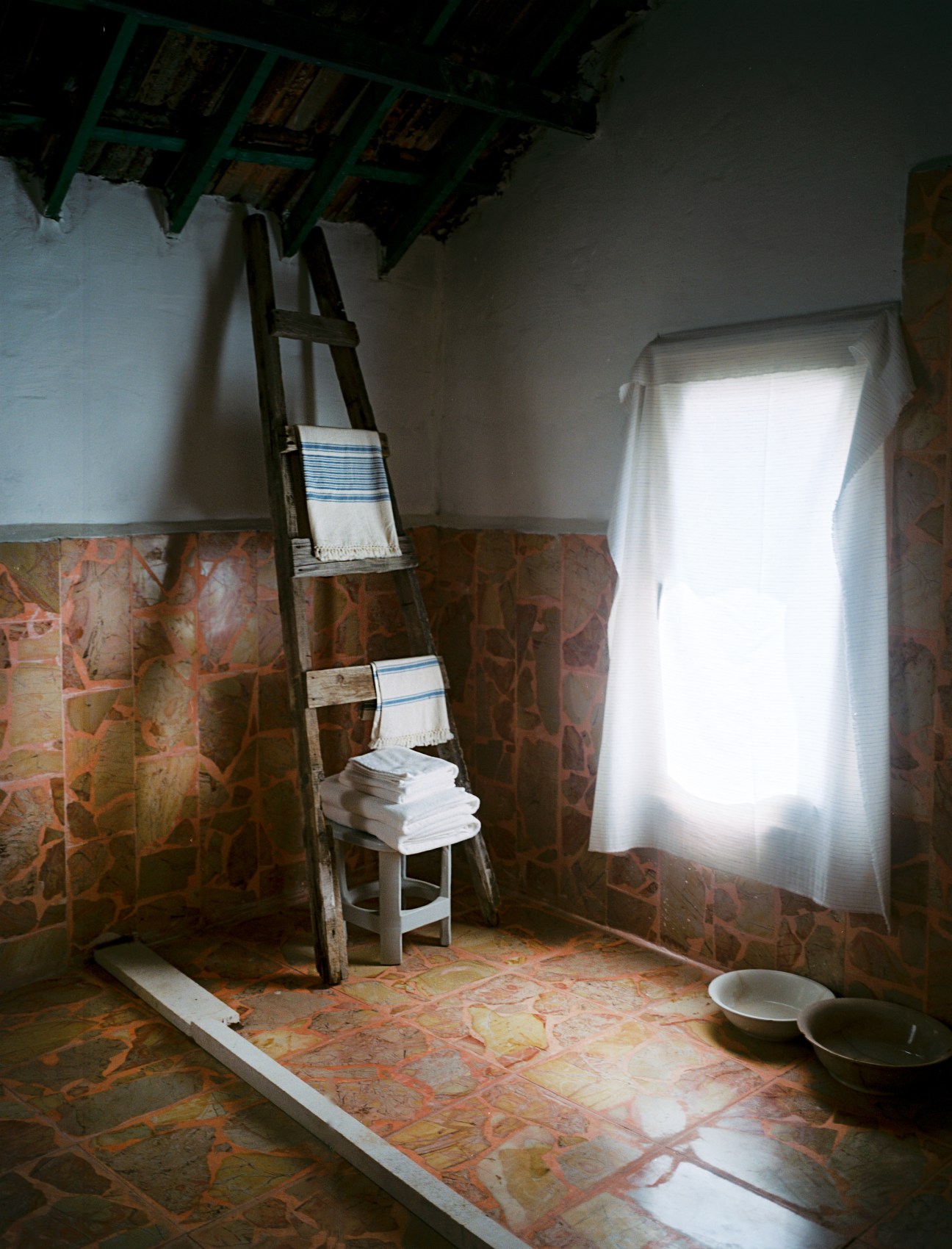 Bathroom interior with red marble floor, wooden ladder with towels, and bright window with white curtains.