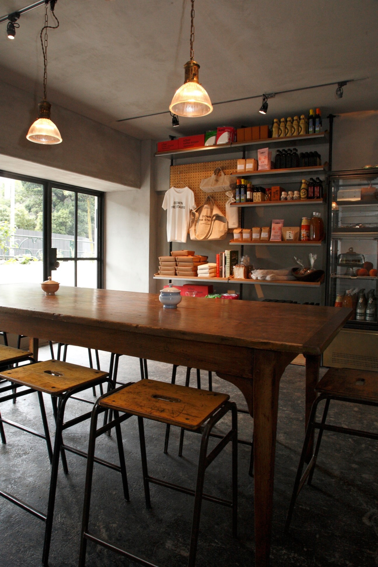 A minimalist retail and dining space with wooden table, hanging pendant lights, and shelving displaying packaged goods and merchandise.