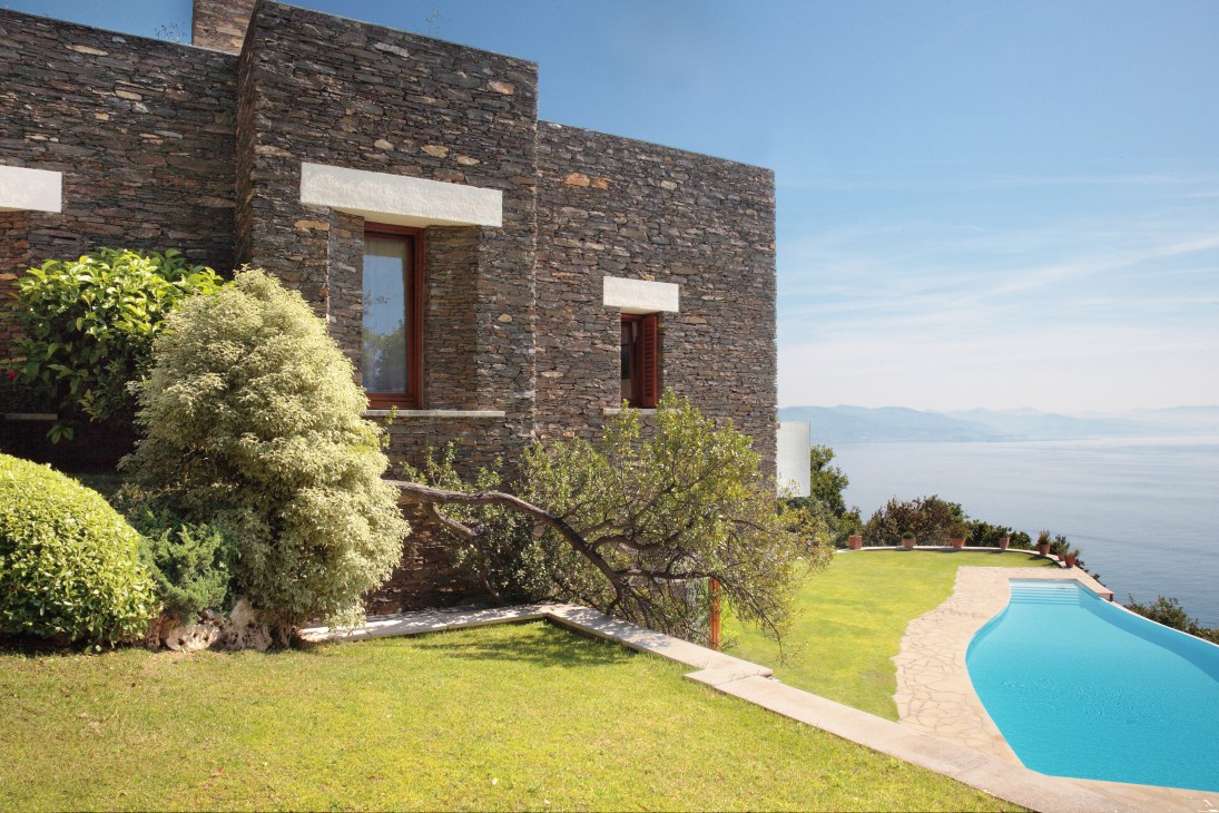 Modern stone villa with clean lines, white-framed windows, manicured lawn and swimming pool overlooking the sea.