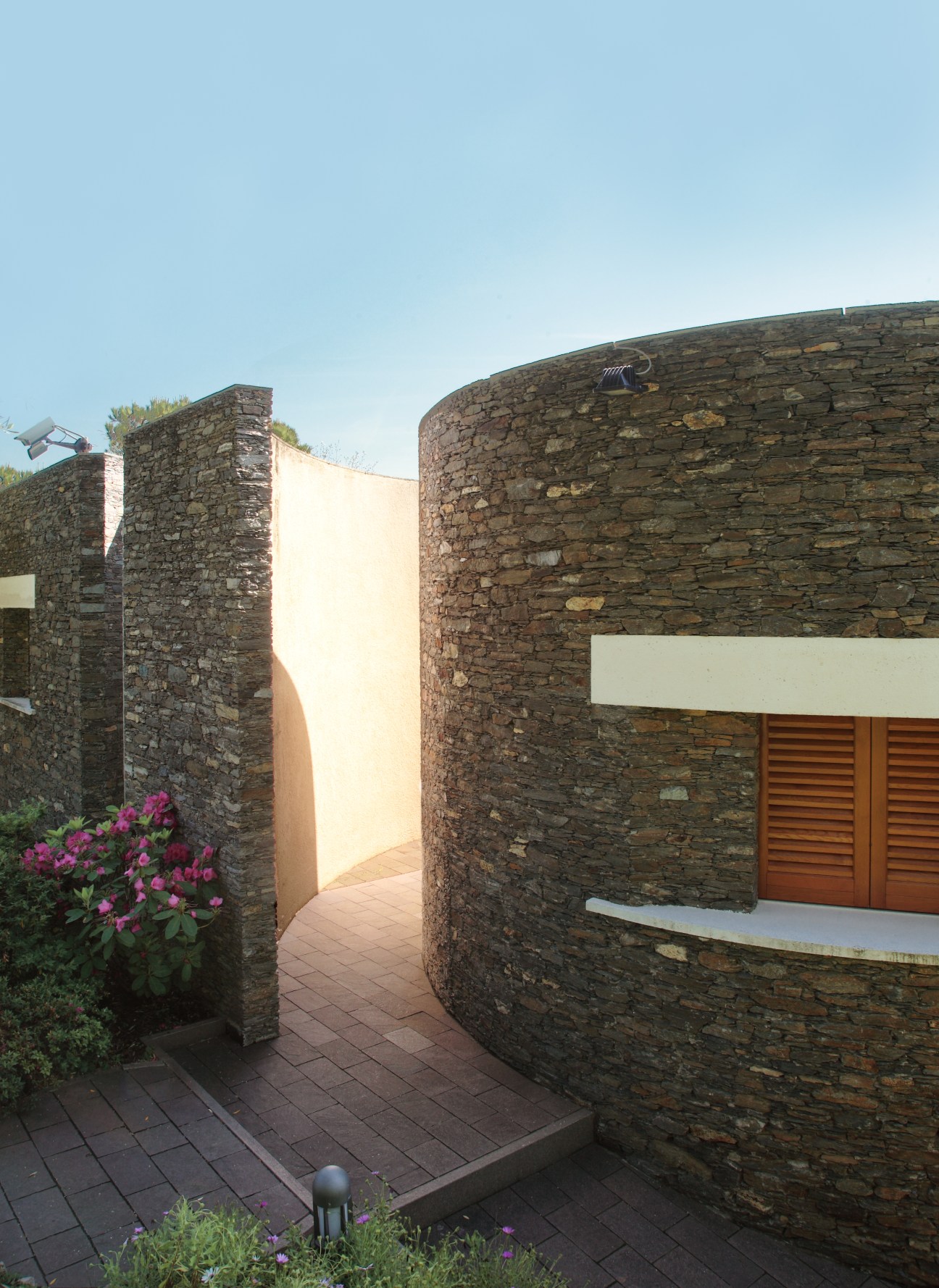 Modern stone villa with curved walls, white accent band, terracotta tile flooring, and magenta flowers.