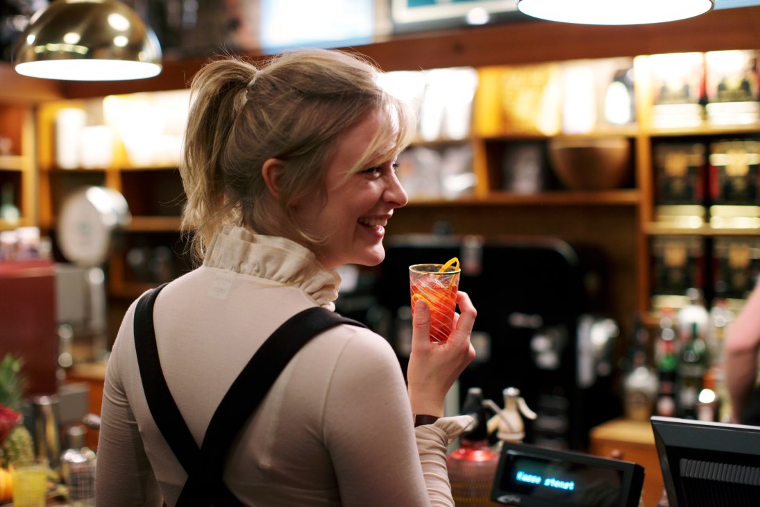 Woman smiling while holding a cocktail at Fuglen café's bar counter with mid-century shelving in background.