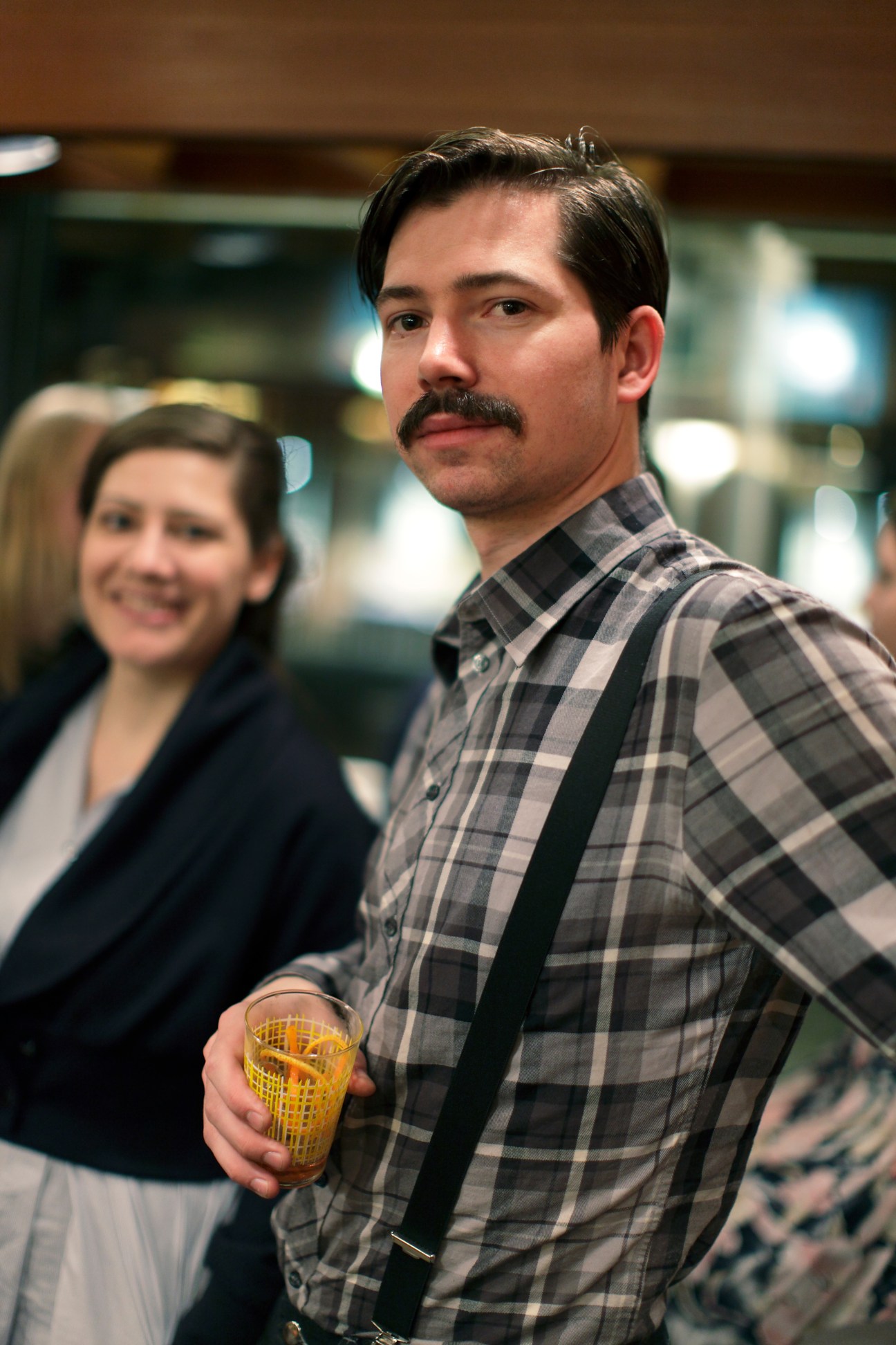 Man with mustache in plaid shirt holding a yellow glass at a cocktail party.