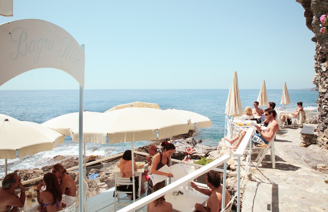 Beachgoers relax under white umbrellas at Bagni Sillo's rocky coastal bathing spot near Genoa, Italy.