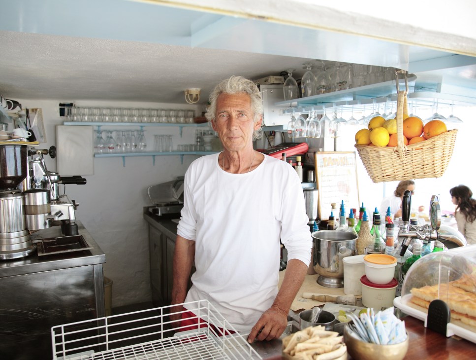 Man in white shirt standing at counter in seaside bathing establishment kitchen with fresh citrus and supplies.