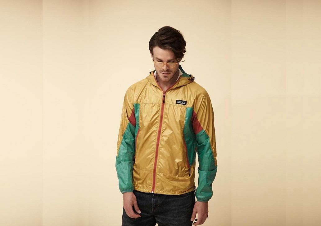 Man wearing yellow and green Manastash windproof jacket with hood against cream background.