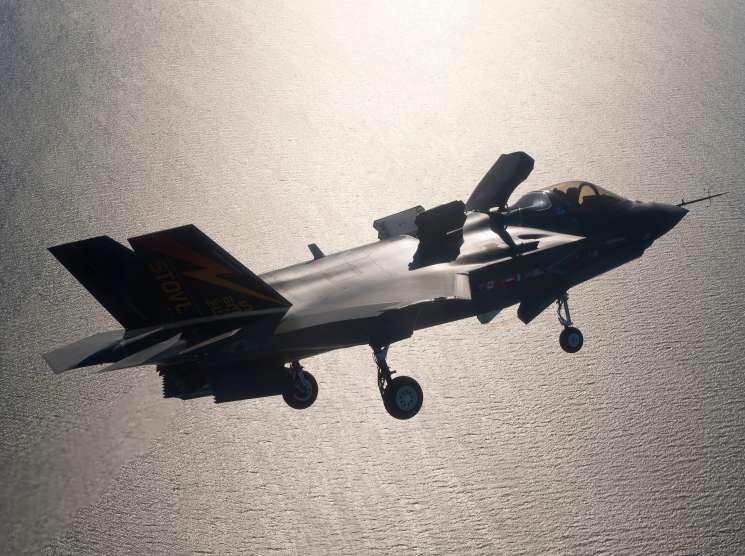 A sleek military stealth fighter jet in flight over water, shown in silhouette against a bright sky.