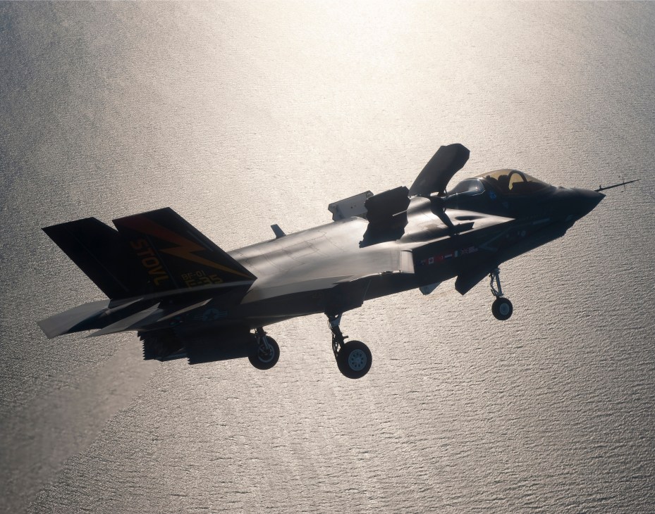 A sleek military stealth fighter jet in flight over water, shown in silhouette against a bright sky.