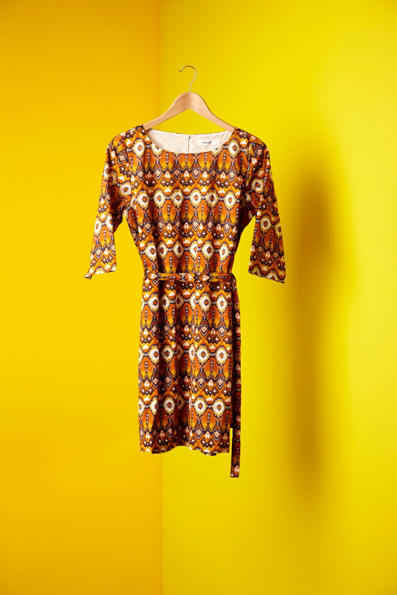 Samuji dress with geometric Moroccan-inspired pattern in orange, brown and cream, displayed on wooden hanger against yellow background.