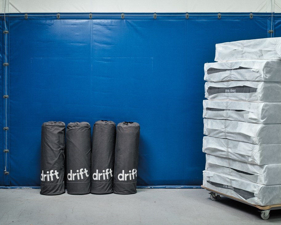 Rolled Drift mattresses in white packaging stacked on a pallet next to compressed black Drift mattress rolls against a bright blue industrial wall.