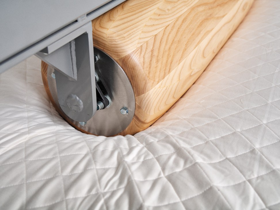 A mattress undergoes durability testing with a mechanical roller pressing down on its wooden base.