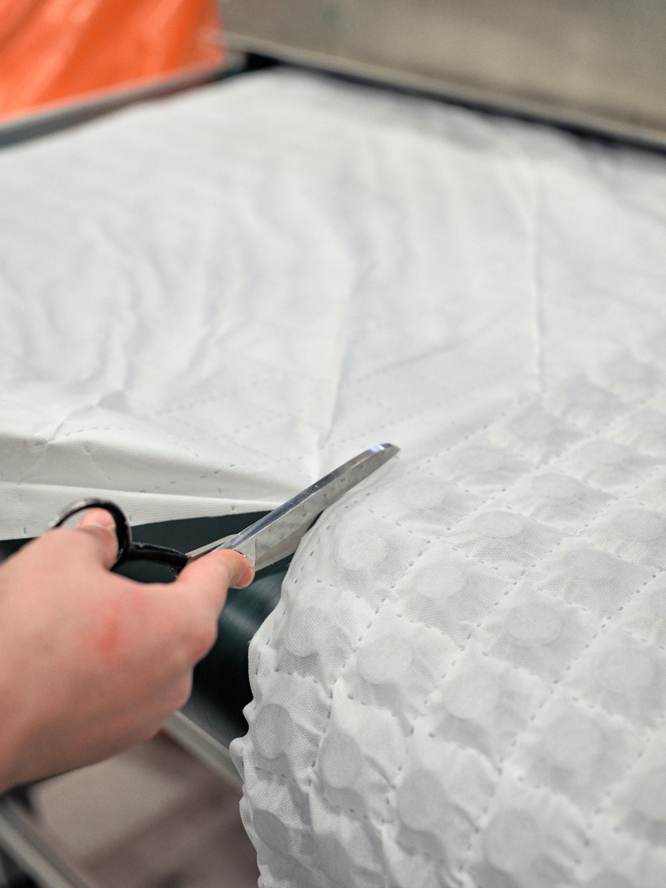 Hand holding a utility knife cutting white quilted mattress fabric during manufacturing.