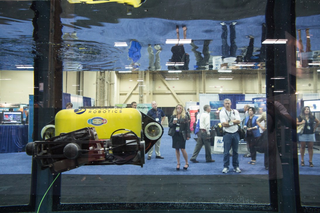 Yellow and black rectangular underwater drone displayed in water tank at trade show with visitors observing behind glass.