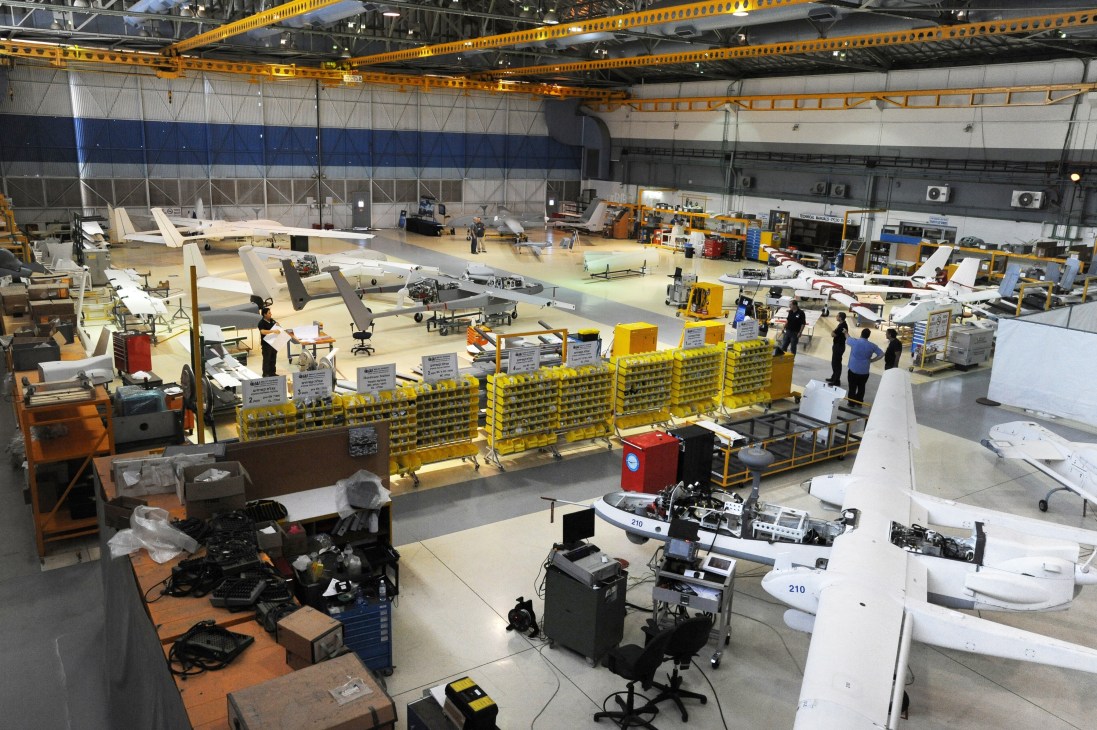 Interior of a large industrial hangar with yellow overhead cranes, containing multiple unmanned aerial vehicles in various stages of assembly and testing.