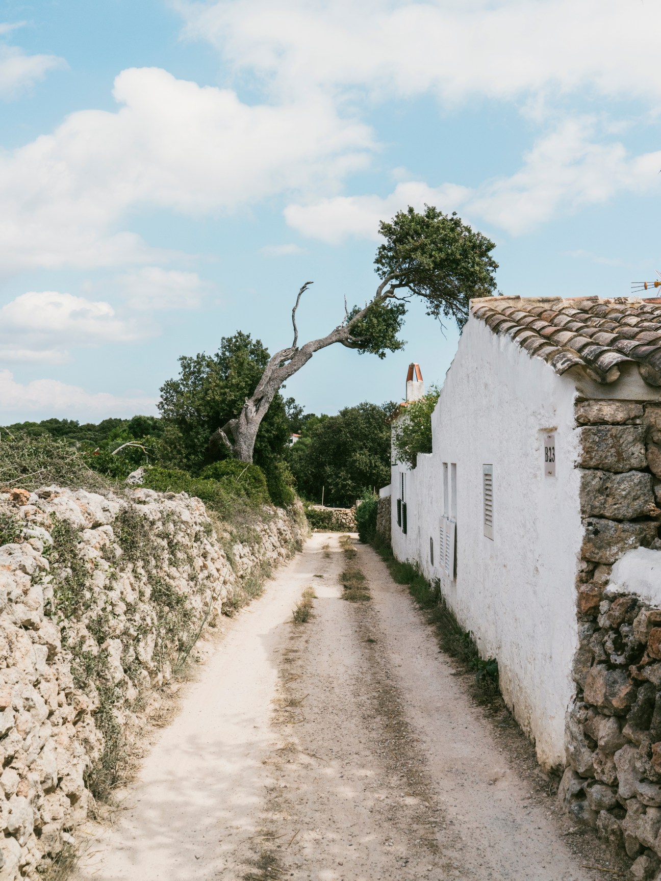 A dirt road lined with white-washed stone buildings and limestone walls under blue sky with clouds in Menorca.